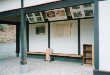 木で出来たベンチや棚がありその後ろの壁に説明書きや資料、上には写真など飾られているトイレ入り口の写真
