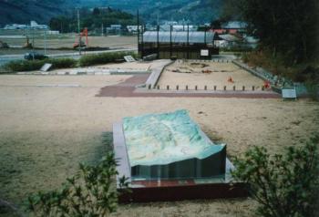 山や畑に囲まれた中にある小さな公園と大きな砂場と茶色の大理石の上に断層の模型が飾られている写真