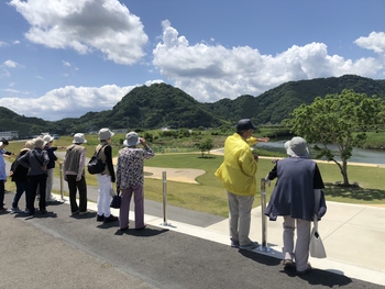 舗装された高台から山の前に広がる芝生と川を眺める人々の写真